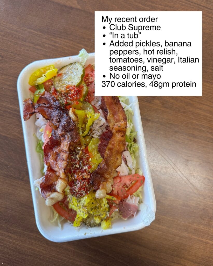 sub in a tub with toppings, vegetables, bacon, and seasonings layered in a bowl-style container